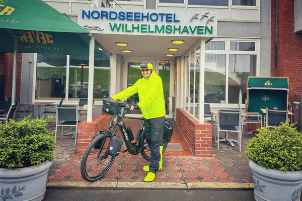 Jens Almer mit seinem Fahrrad in Regenkleidung vor einem Hotel in Wilhelmshaven